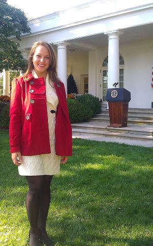 Marcelle in a red coat and smiling as she stands in front of the empty presidential podium in the rose garden