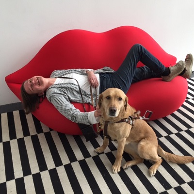 Beth lounging on a ruby red couch shaped like lips, with Seeing eye dog Whitney standing in front of the couch. 
