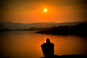 A man sitting on the bank of a body of water watching a very vivid, orange-hued sunset