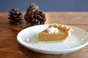A piece of pumpkin pie on a wooden table with two pinecones in the background