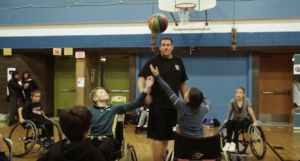 Students in a gym testing out wheelchairs and tossing up a basketball