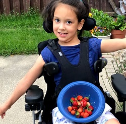 A young girl in a wheelchair, arms spread wide, with a bowl of strawberries in her lap