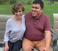 Ali, a young woman with short brown and wavy hair smiling with boyfriend Juan, turned slightly towards her, also smiling. They are sitting on a bench and each has an arm around one another.