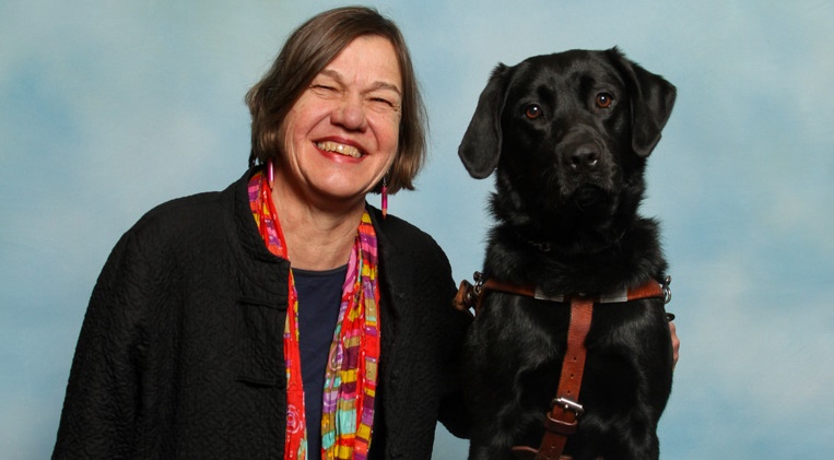 Beth wearing a black shirt with a colorful scarf, standing next to her service dog, a black lab. 