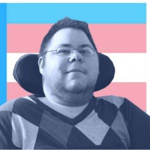 Dominick, a man using a wheelchair and wearing a pair of glasses smiles against a backdrop of an LGBTQ/Trans flag