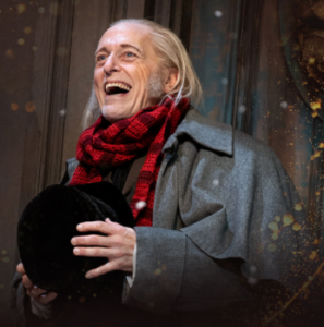 Actor Larry Yando in the role of Ebenezer Scrooge, holding a hat, wearing a grey coat and scarf, smiling and laughing