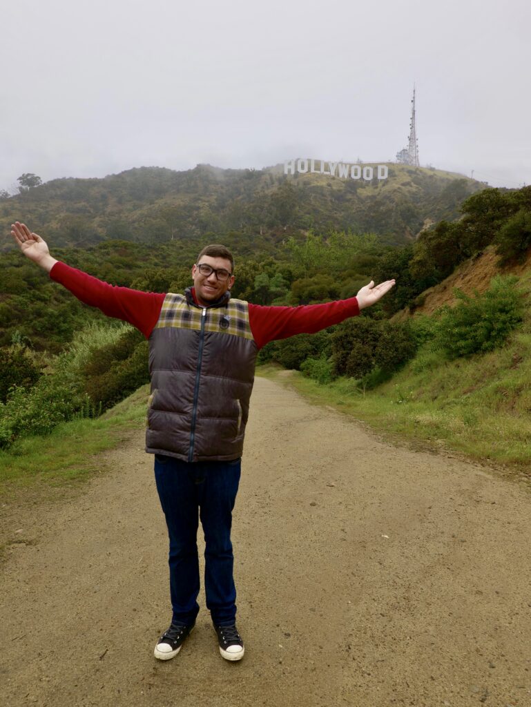 Scott Outside wearing a vest, arms outstretched, in front of the iconic Hollywood sign.