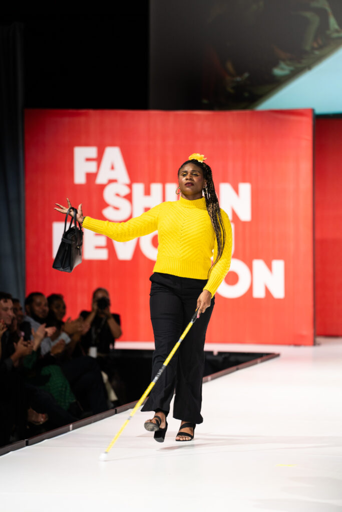 A woman using a yellow cane and wearing a yellow top and black pants on the runway 