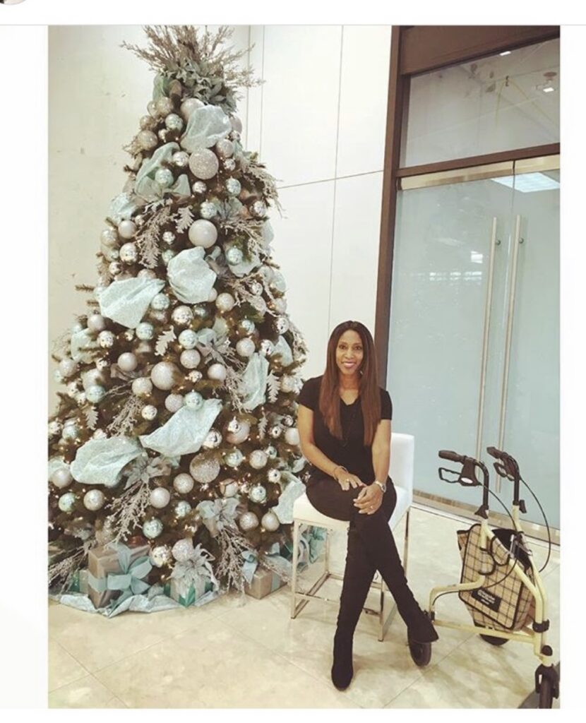 A black woman wearing a black shirt and pants is sitting on a white chair in front of a tall Christmas Tree decorated with Robin's Egg Blue ornaments and decor.