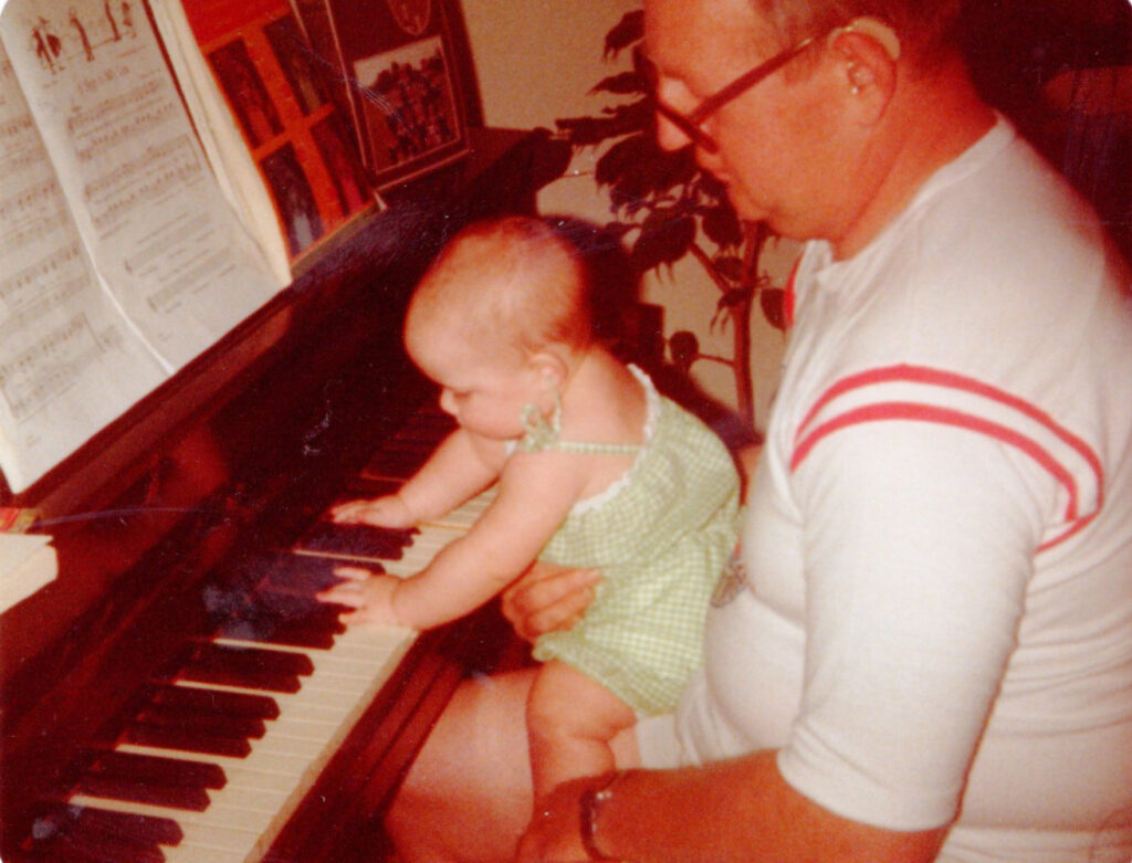 Photo from the 70s. Man with a hearing aid holding a baby, sitting by a piano