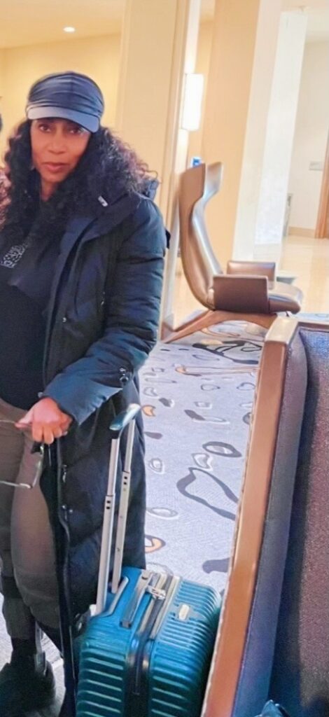 A Black woman using a black cane with a black baseball cap covering her dark brown shoulder-length curly hair is posing by a blue suitcase at a hotel, waiting for her transportation to the airport.