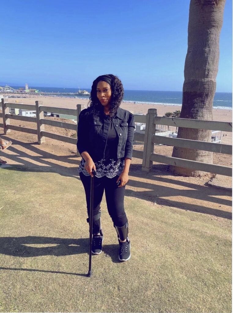A Black woman with brown curly hair blowing in the wind and a black headband around her hair is wearing a black cane and black leg braces with all back attire on, including a denim jacket blouse and tennis shoes/sneakers. She has white decorative designs on her blouse.