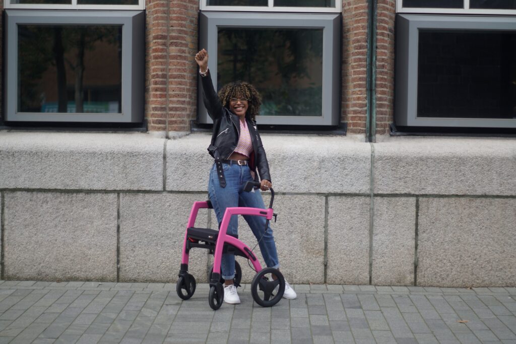 A Black woman wearing glasses and using a pink rollator with black wheels stands with one arm on the rollator and the other raised in the air. She has a curly natural hairstyle with blonde highlights, and she is wearing a short waist-length leather black jacket with a pink midriff blouse, blouse, jeans, and white tennis shoes.