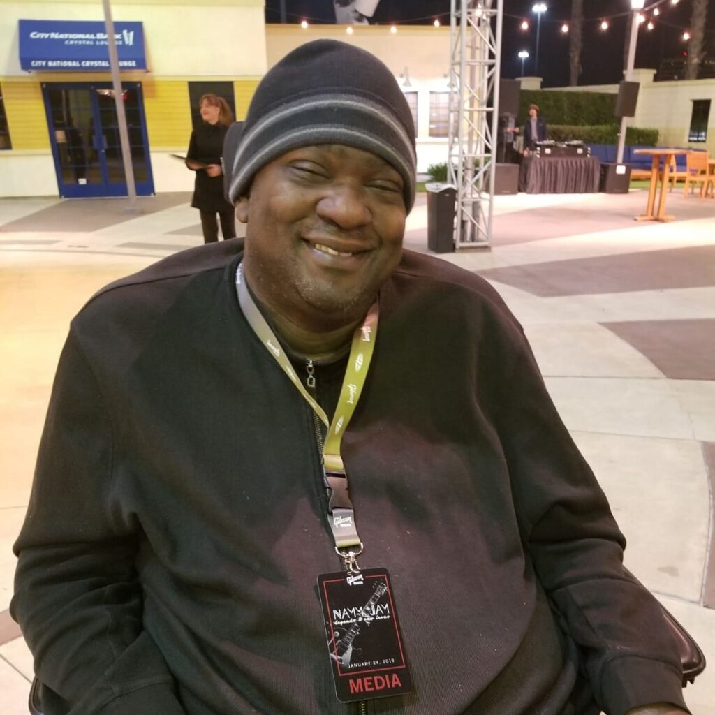 Paul wearing a beanie and a lanyard around his neck. He is using his wheelchair at a convention