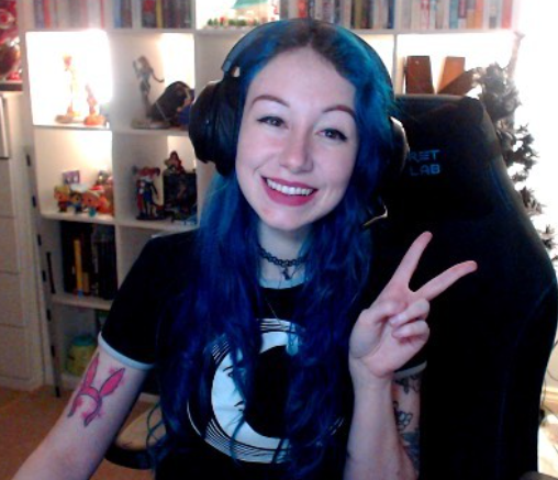 Mollie, with long blue hair sits in a gaming chair, and they are giving the peace sign