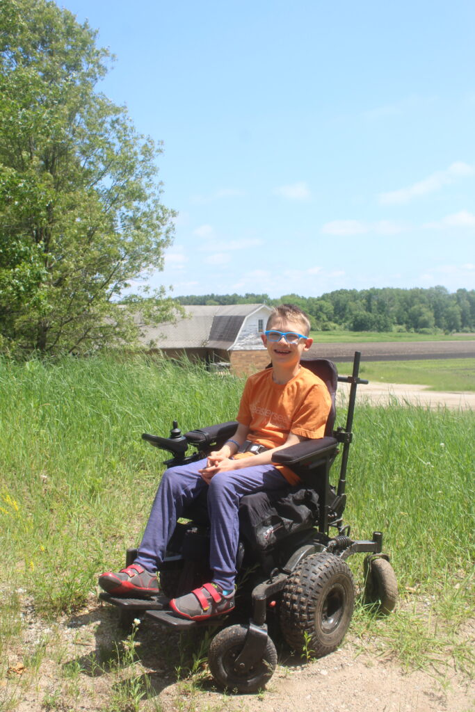 Evan, a 12 year old boy, is using his all-terrain wheelchair outside on the farm. He has blue glasses and an orange shirt with jeans