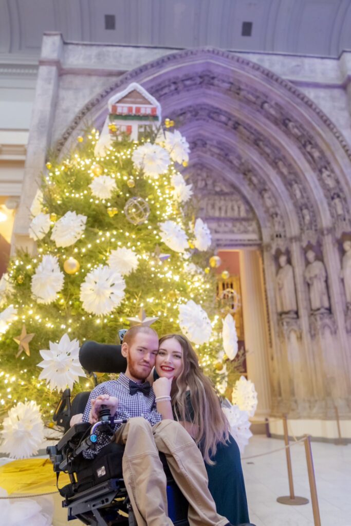 Grant, using his wheelchair, is smiling next to his girlfriend who leans into him. They are in front of a tree with lights