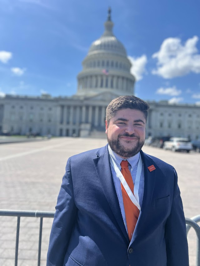 Matt wearing a blue suit with an orange tie, standing in front of the US Capitol building. Blue sky and clouds.