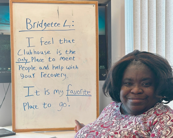 Bridgette, a woman stands in front of a white board that says "I feel the clubhouse is the only place to meet people and help with your recovery. It is my favorite place to go!