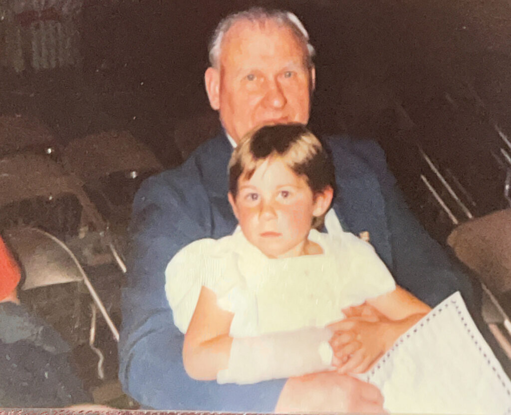 Dom as a young child, wearing a white dress and an arm brace. They are sitting on their dad's lap