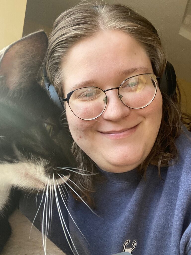 Megan, wearing round metal glasses is smiling and hugs their cat