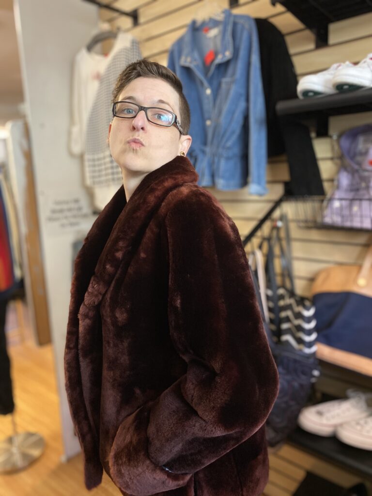 Raven at a thrift store wearing a fur jacket, making a silly kissy face at the camera 