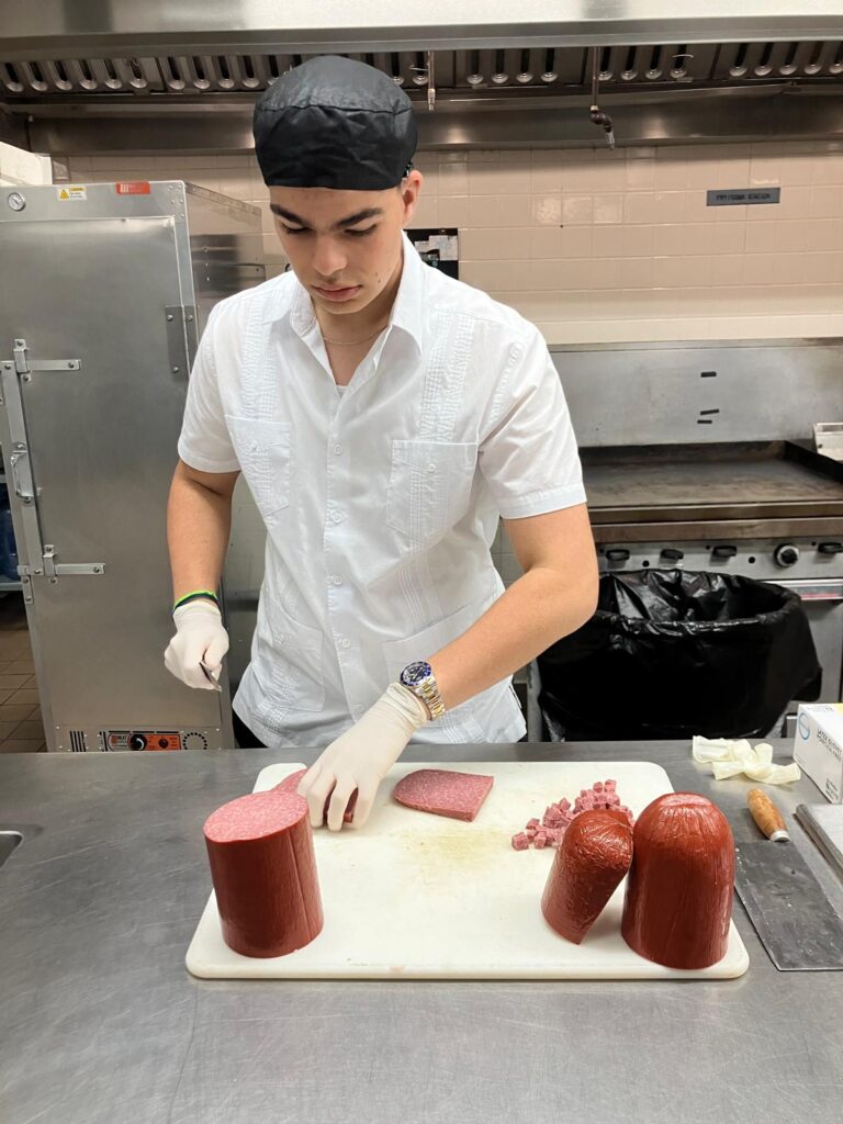 Sebastian in an industrial kitchen, cutting up sausages, wearing a chef hat