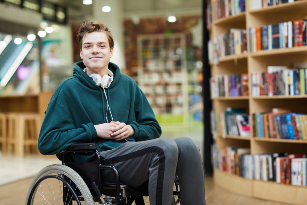 Disabled student with headphones on neck siting in wheelchair and looking at camera in modern library or bookstore