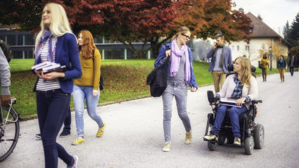 Two friends, one using a wheelchair, walking together through the park, other students are walking around.