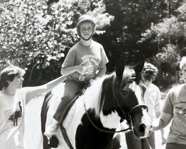 A black and white photo of Rhonda wearing an Easterseals Camp shirt, riding a horse, with two volunteers beside her 