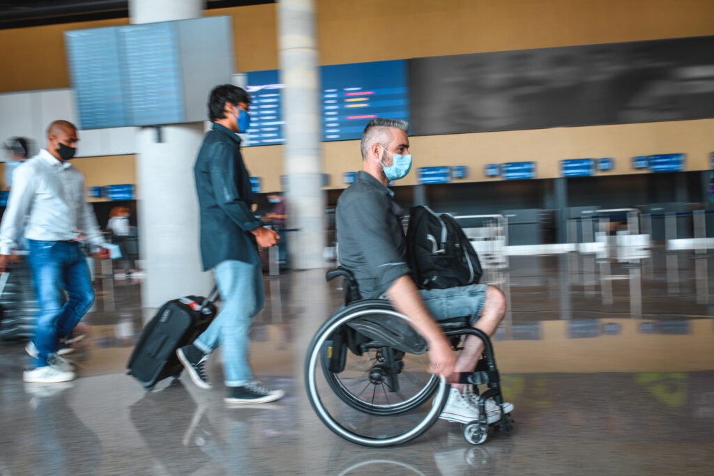 Diverse people on the move at the airport. Wearing protective face masks during pandemic. Blurred motion of a wheelchair user.