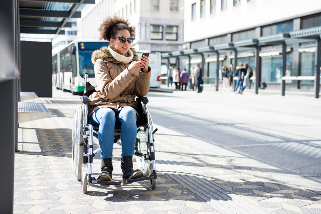 Disabled woman in a wheelchair waiting at a bus station and using smart phone