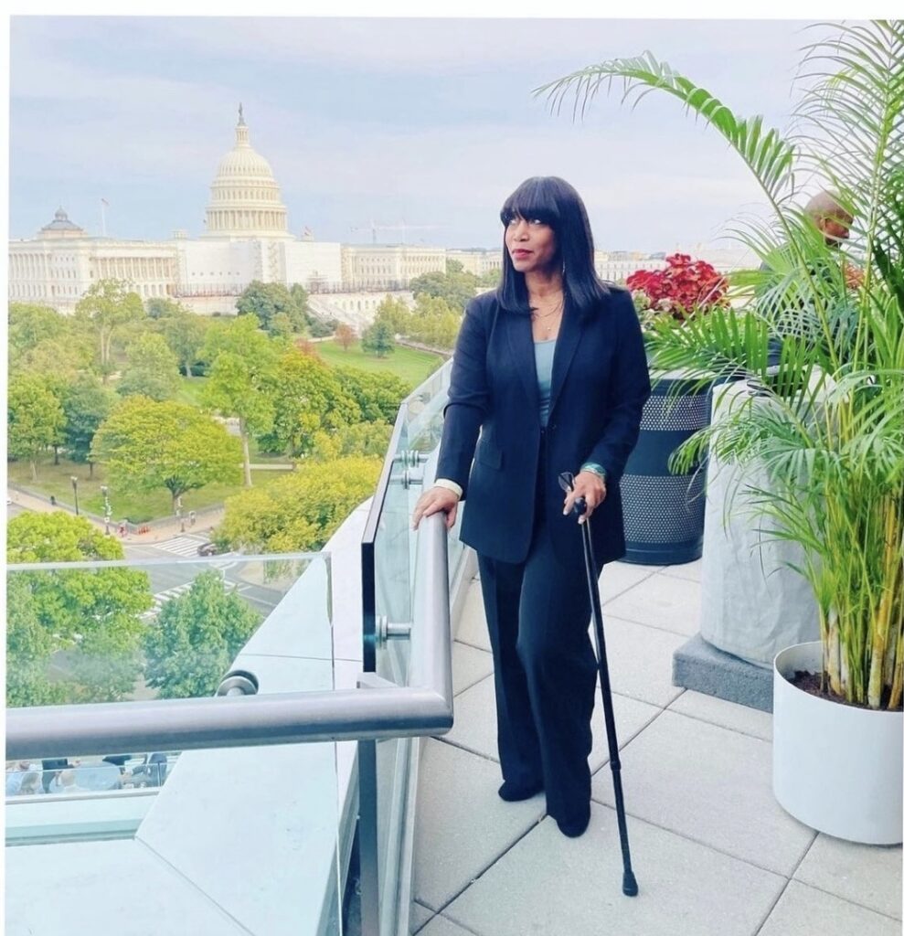 Andrea, a Black woman using a cane, stands on a balcony overlooking the US Capitol 