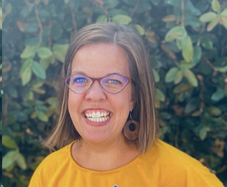 Leah smiling, plants behind her. She is wearing a yellow top, red glasses. and is smiling