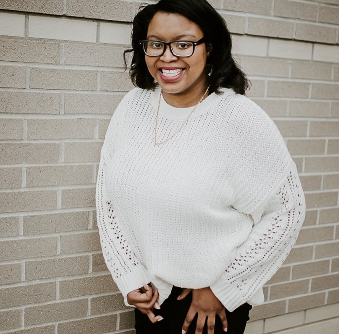 Keah, a Black woman wearing a white sweater, smiles while standing against a wall.